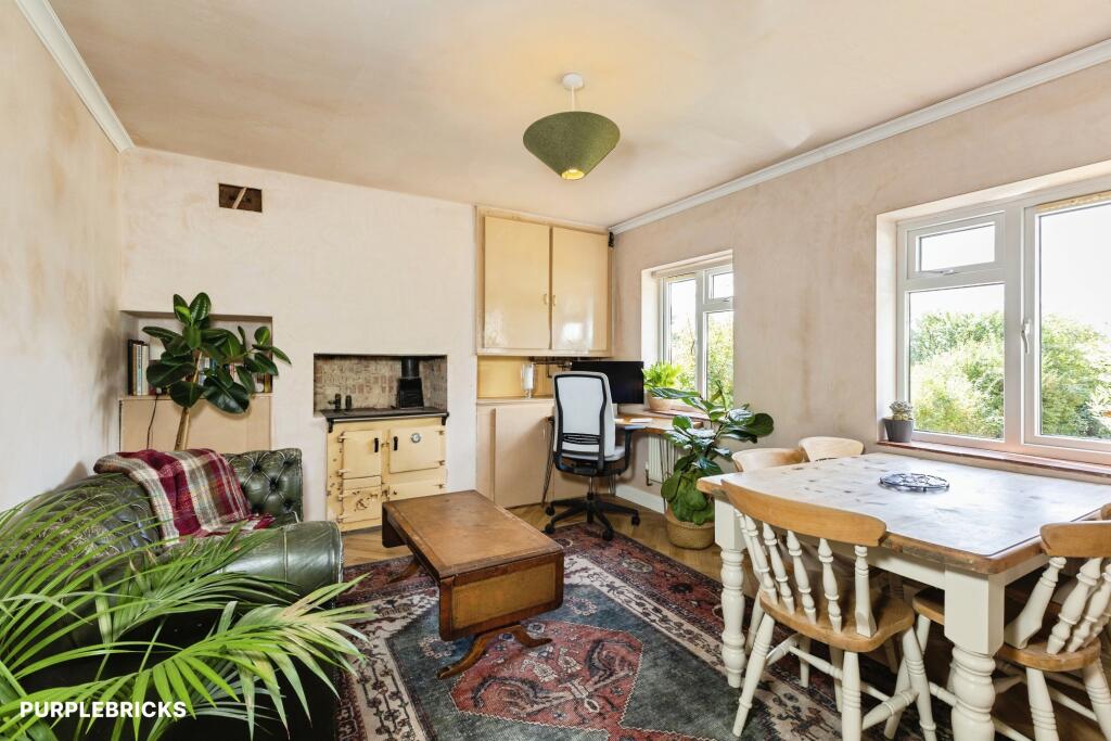 Interior multi-purpose room with bedroom area, kitchenette, dining table, and workspace, featuring wooden furniture, potted plants, and large windows with natural light.