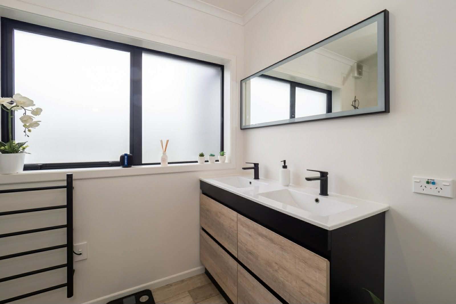 Modern bathroom interior with dual vanity sinks, black framed mirror, and large window with black frames overlooking the exterior.