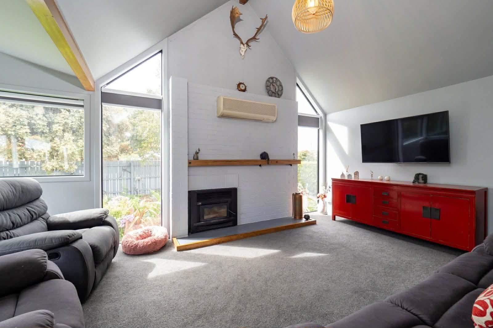 Interior living room with vaulted ceiling, fireplace, wall-mounted TV, red credenza, black recliners, and glass doors opening to a deck with forest views.