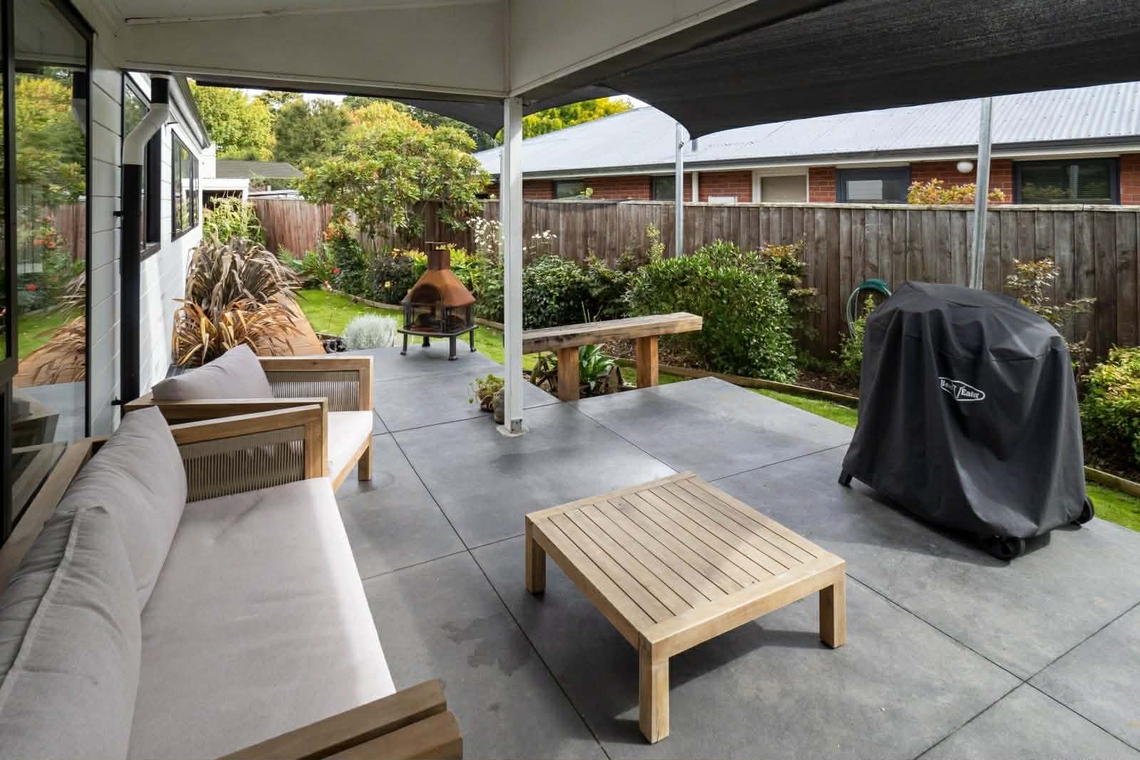 Exterior covered patio with modern outdoor furniture, concrete flooring, and views of a manicured garden with wooden fence and brick homes beyond.