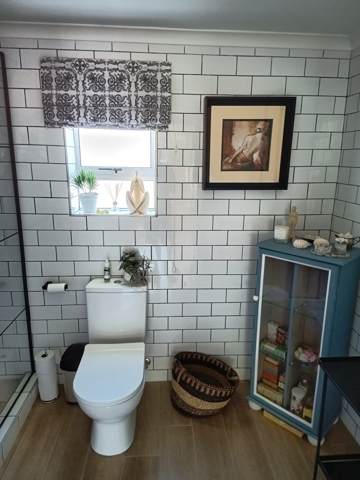 Interior bathroom with white subway tile walls, toilet, pedestal sink, window with decorative roman shade, framed artwork, blue storage cabinet, and wooden flooring.