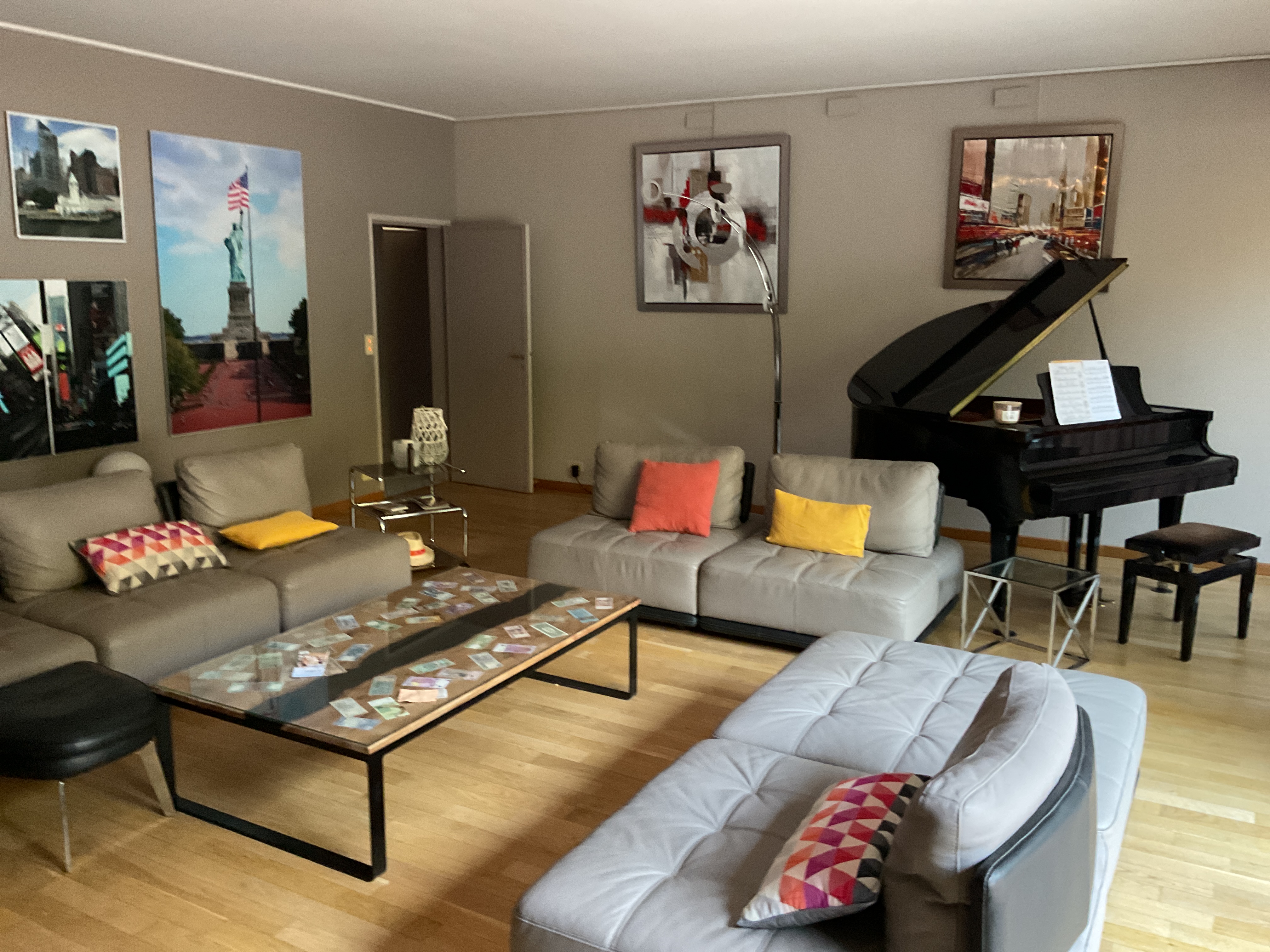Interior living room with multiple gray sofas, a grand piano, wooden floors, decorative artwork on walls, and colorful accent pillows.
