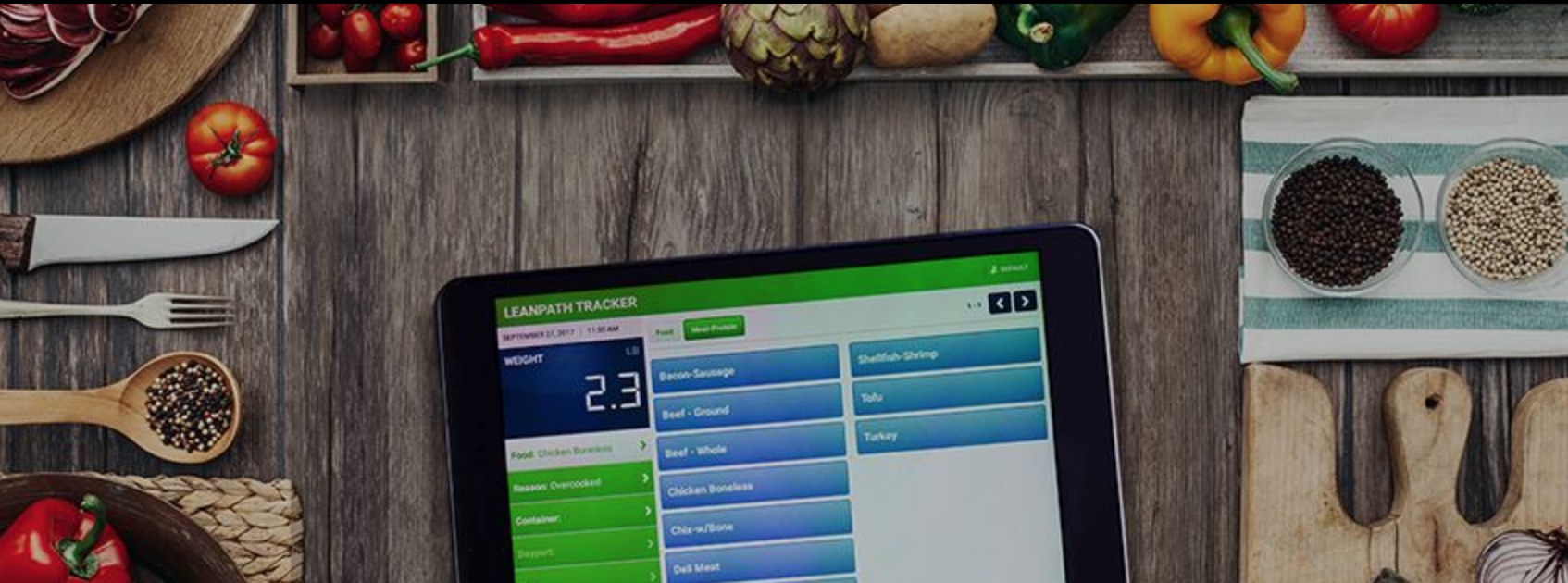 The Leanpath Tracker touchscreen interface on a tablet showing a food-waste weight of 2.3 lb, surrounded by fresh ingredients on a wooden counter