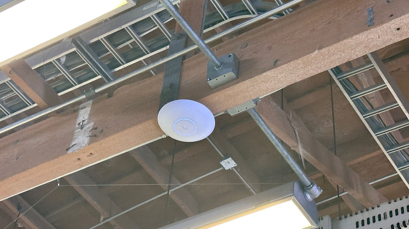 Round UniFi access point installed on an open industrial ceiling near overhead LED linear lights