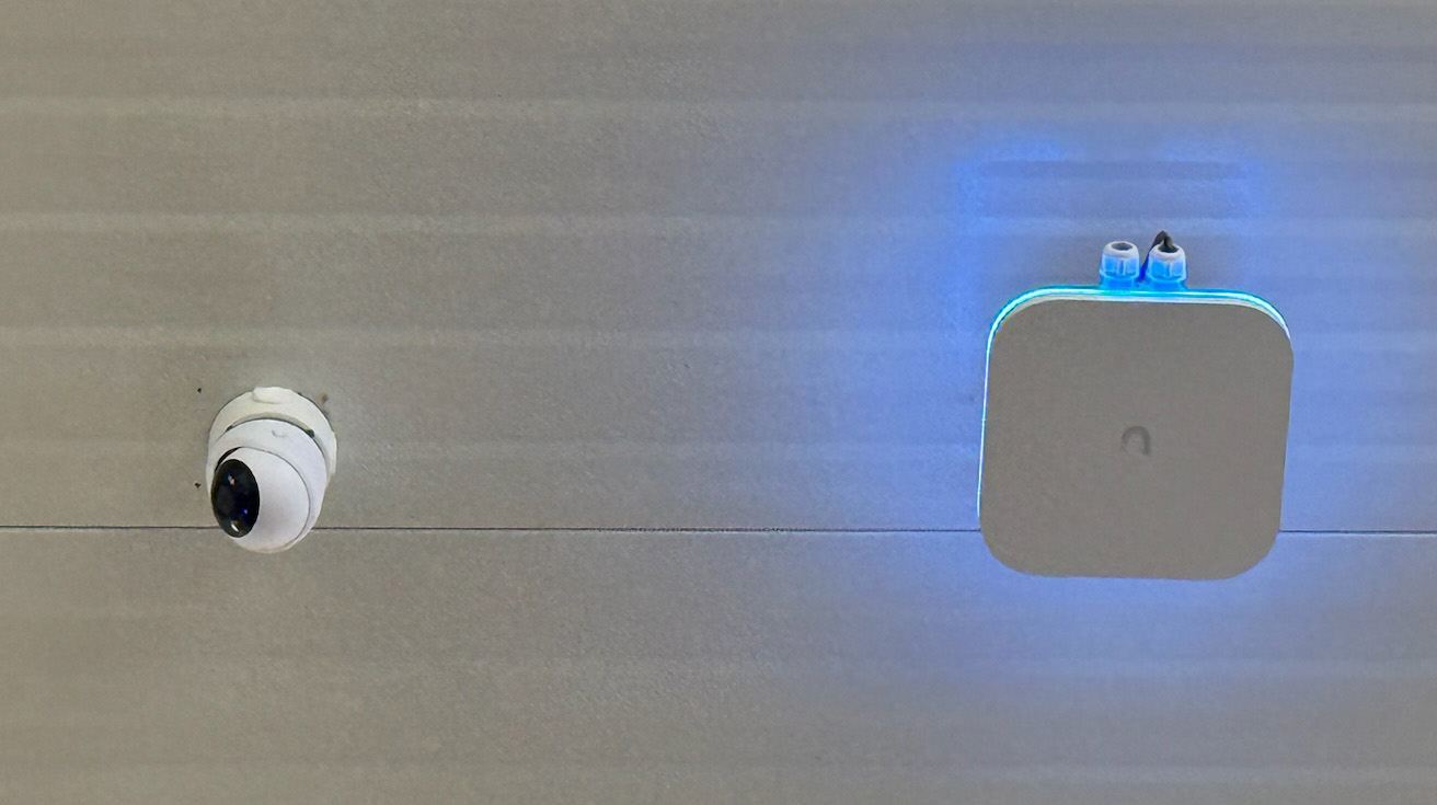 Square UniFi access point mounted externally on a light colored building wall with blue backlight turned on