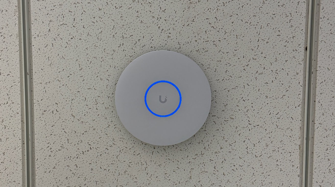 Round UniFi access point installed on an open industrial ceiling near overhead LED linear lights