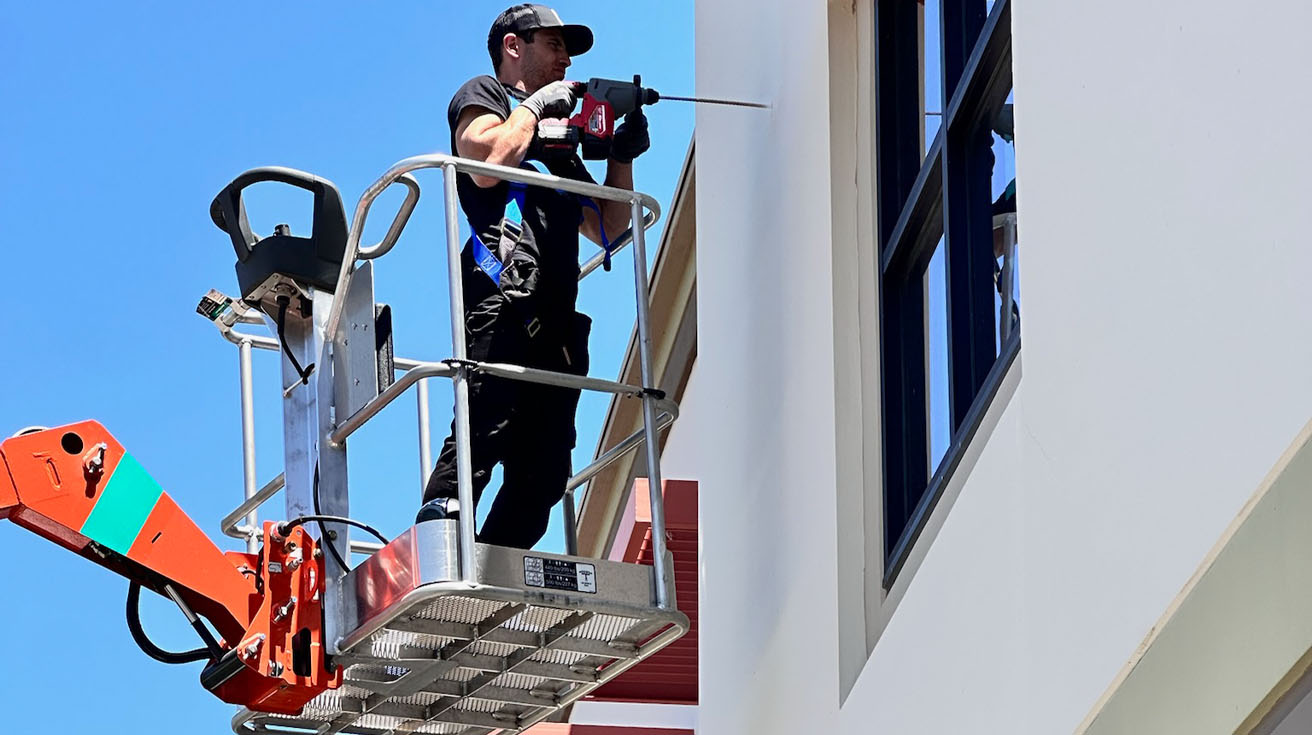 a certified field engineer in a boom lift drilling into a commercial building exterior for physical security infrastructure installation.