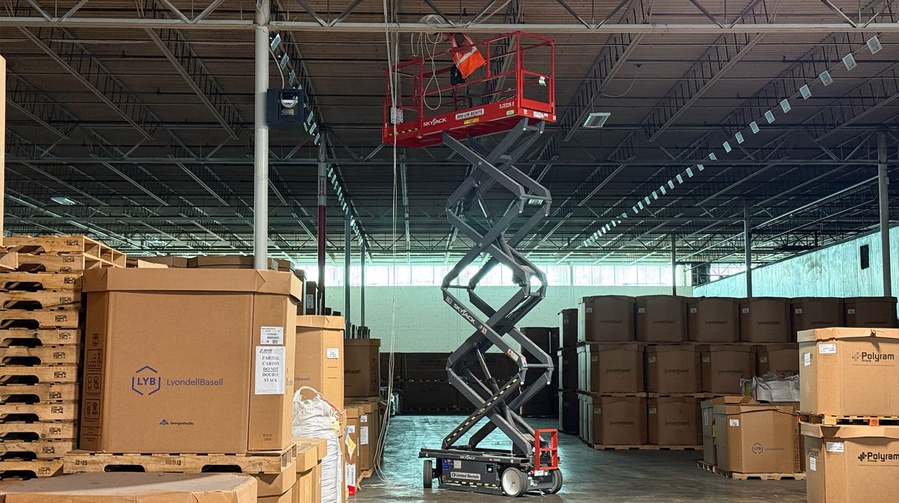 A technician operating a scissor lift to install high-density network infrastructure in a logistics warehouse.