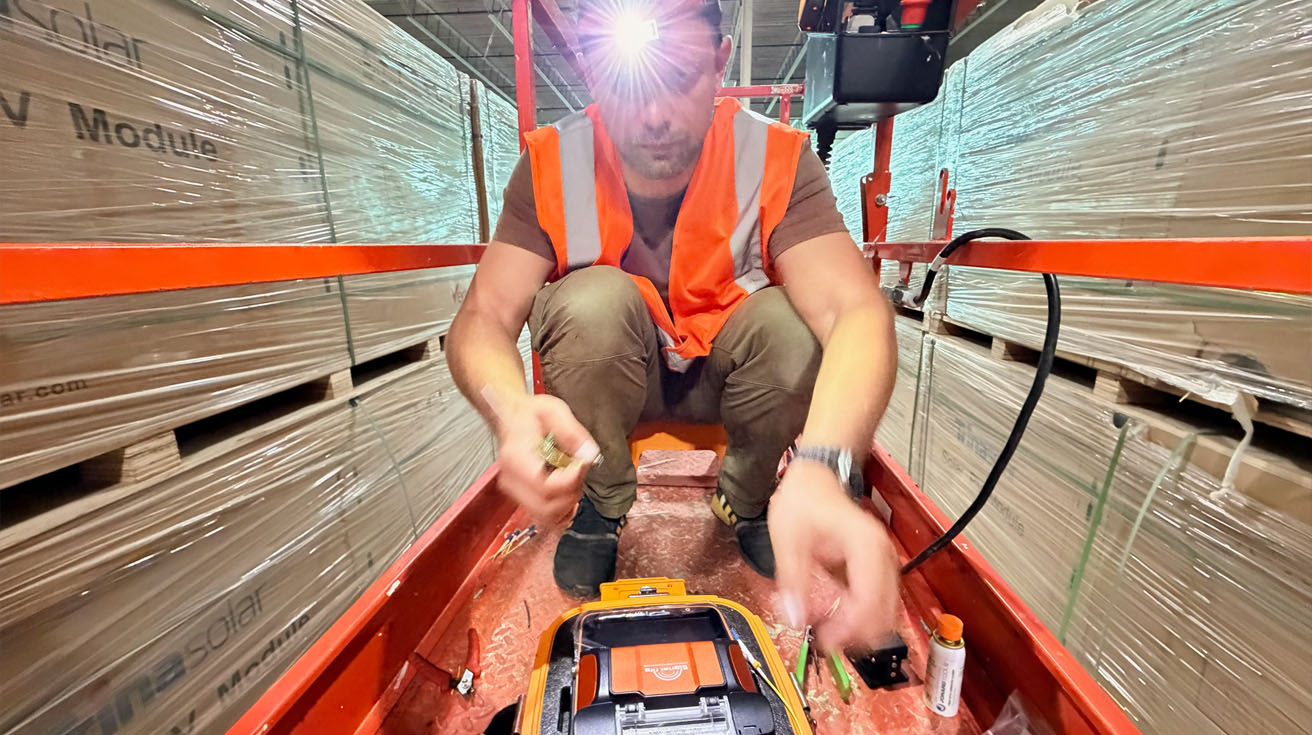 A network engineer operating a fiber optic fusion splicer on a scissor lift inside a large commercial warehouse.
