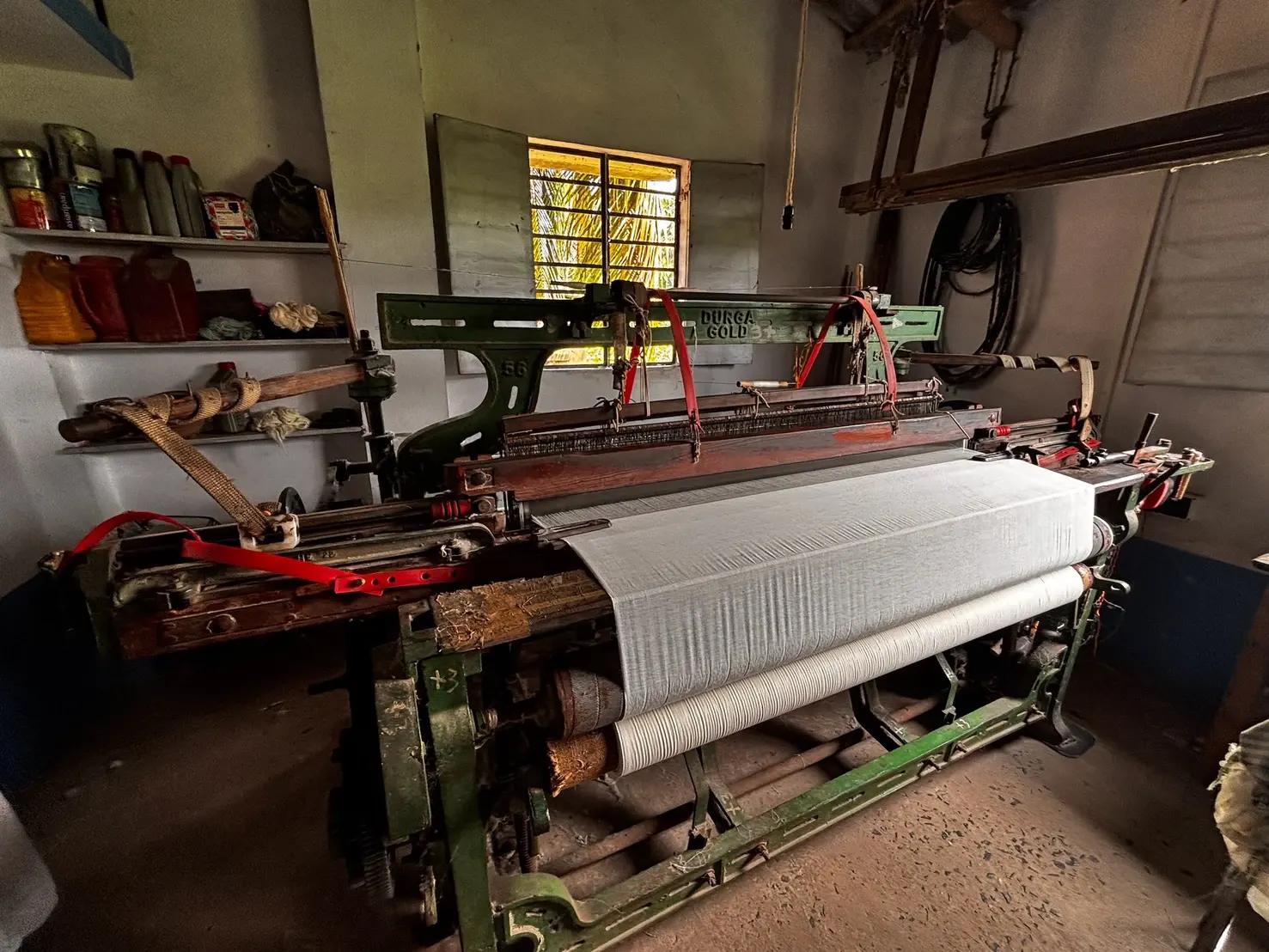 A power loom set up in a workshop, white warp threads stretched across it as cloth comes off the beam