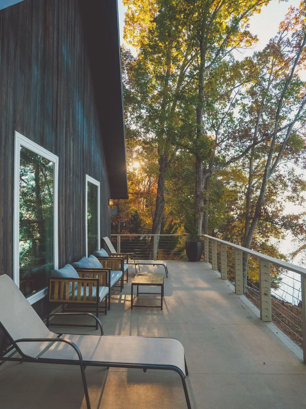 A long deck with cable railings and chairs outside a dark-clad house at sunset