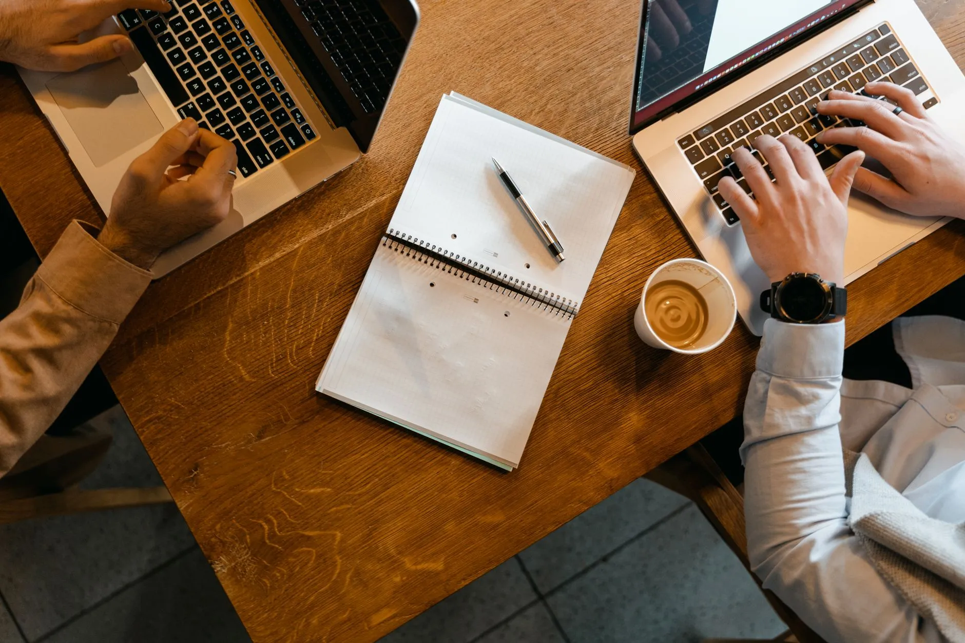 two people typing on laptops side by side at a wooden table, seen from above
