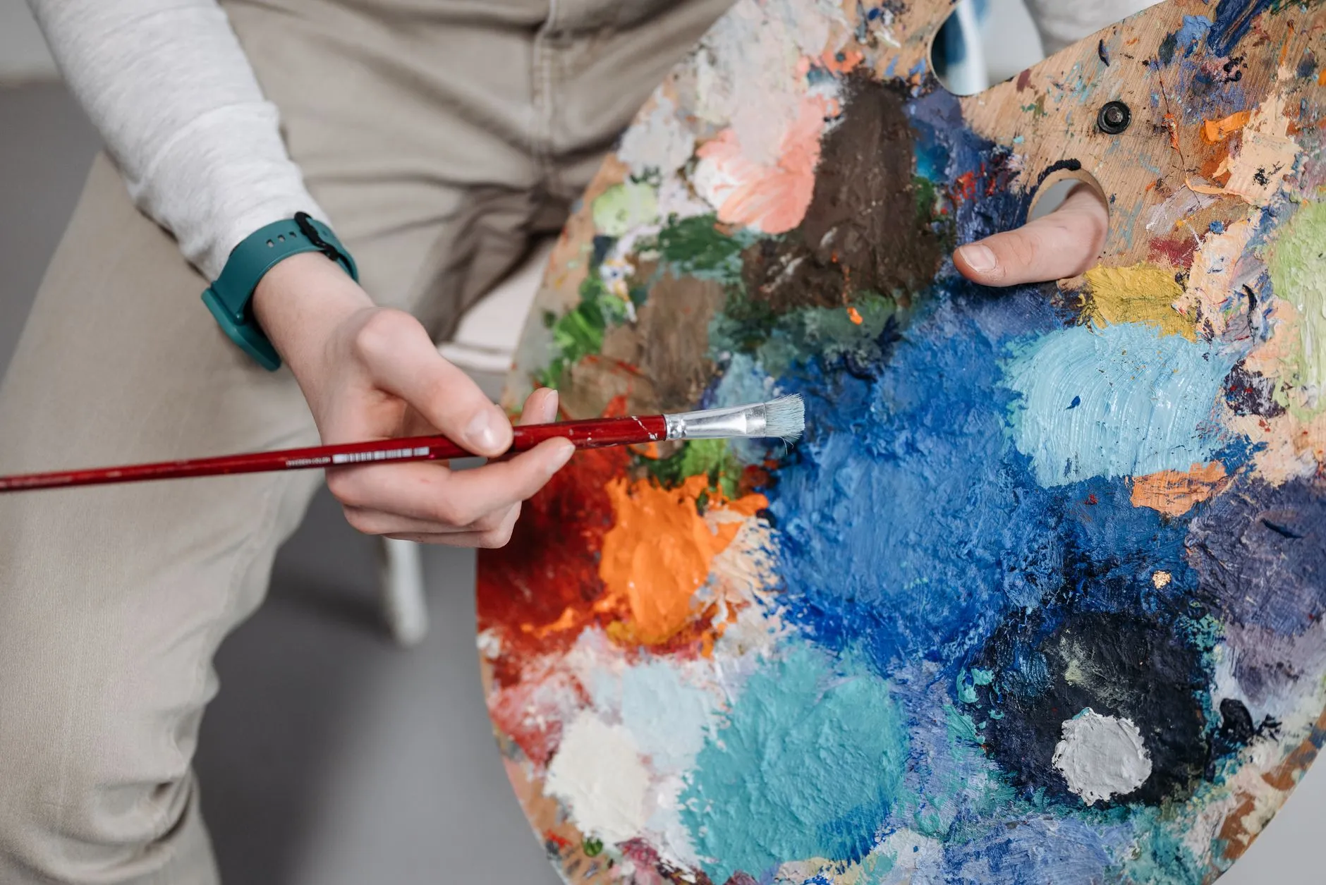 a hand holding a paintbrush over a palette of bright mixed paint