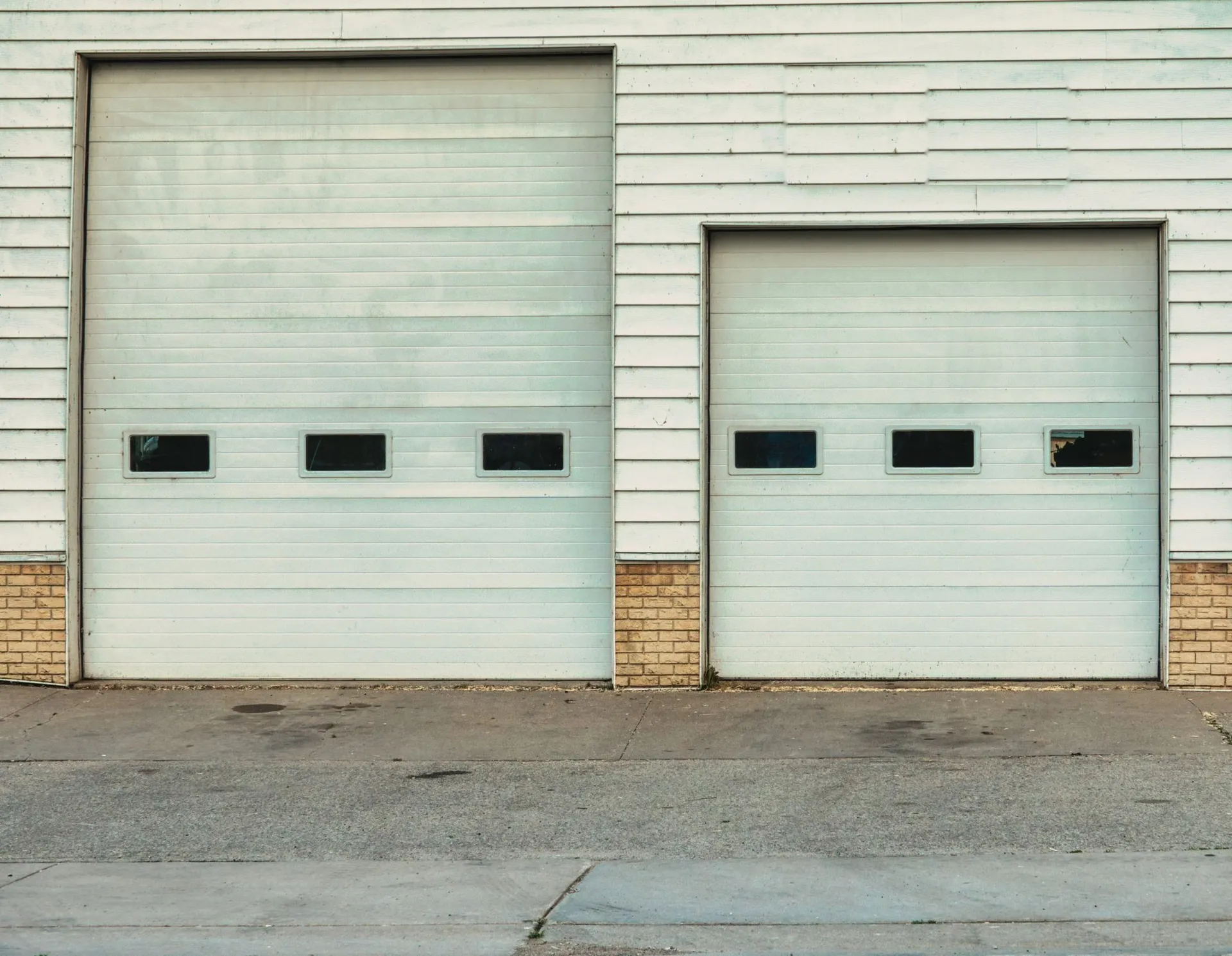 a white garage with two roll-up bay doors and a brick base, facing an empty concrete apron