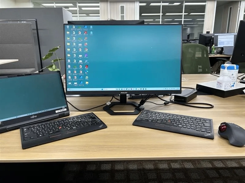 オフィスのデスクにノートPC、外付けモニター、キーボード、トラックボールマウスが置かれている