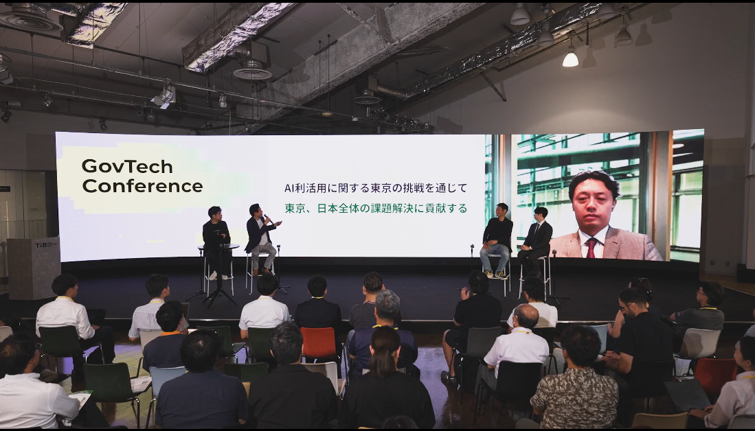 「GovTech Conference」のステージで、5人の登壇者がディスカッションを行っている様子。登壇者の背後のスクリーンには「AI活用に関する東京の挑戦を通じて 東京、日本全体の課題解決に貢献する」というテーマとオンライン参加した松本氏の顔が表示されている。観客が着席して聴講している。
